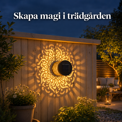 Närbild på en svart solarcellslampa i metall monterad på en ljus vägg som sprider ett detaljerat mandalamönster av ljus.