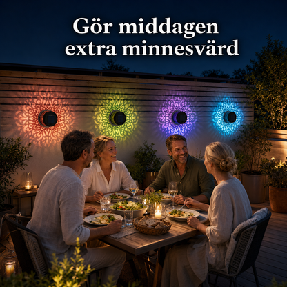 En middag på altanen på kvällen med vänner runt ett bord. Solarcellslampor på trästaketet kastar vackra och färgstarka mandalamönster i bakgrunden.