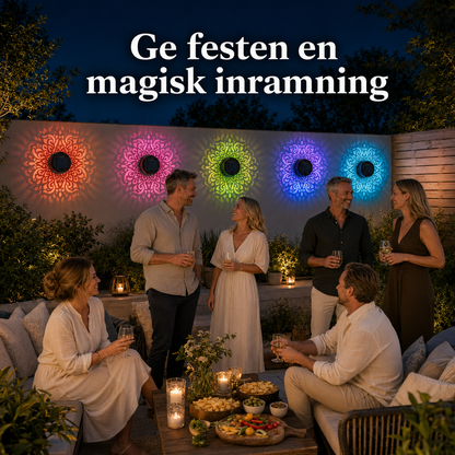 En trädgårdsfest på kvällen där en grupp vänner står och pratar på en uteplats. På väggen bakom lyser flera solarcellslampor i olika färger med mandalamönster.